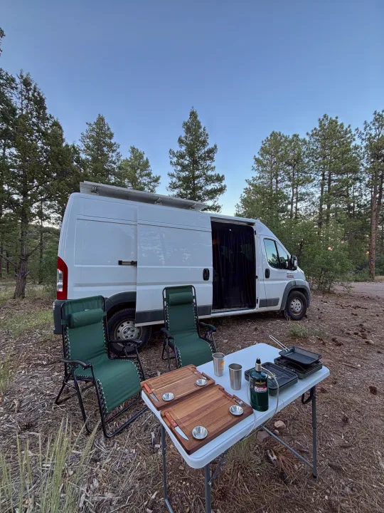 RV rental: Camper van in a forest, perfect for van life adventures, with chairs and outdoor cooking setup.. RAM Promaster 2500 2018