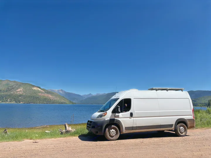 White RV rental van by a blue lake & green mountains under a clear sky. A dog peeks from the window, enjoying the scenic view.. RAM Promaster 2500 2018