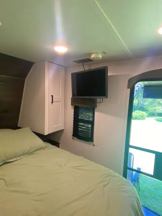 Inside an RV rental bedroom: light green bed, wall-mounted TV, white cabinets, and a window with blinds overlooking greenery.. Keystone RV Hideout 2022