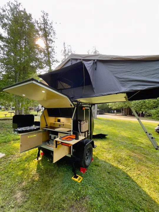 An open compact rv rental trailer with a rooftop tent and outdoor kitchen setup in a sunny wooded campground.. Nutshell Campers Nutshell Camper 2025
