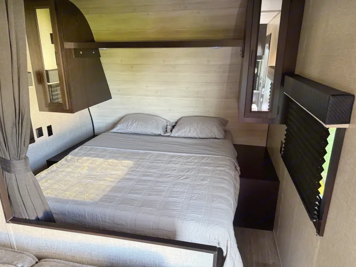 Inside an RV rental: a neatly made bedroom with a light gray bed, overhead storage, and window with blinds.. Jayco Jay Flight SLX 2022