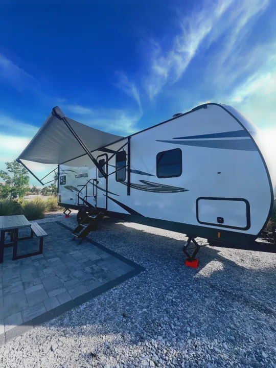 This rv rental is set up at a sunny campsite with an extended awning, picnic table, and paved patio, ready for adventure.. SportTrek Venture 2024
