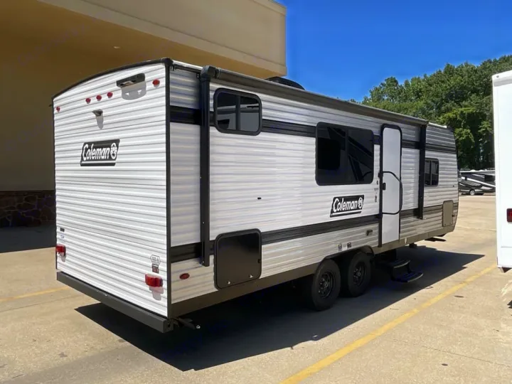 Rear-side view of a Coleman travel trailer, ideal for RV rental. Features a light-colored exterior, dual axles, windows, and entry door.. Coleman 23B Keystone 2025