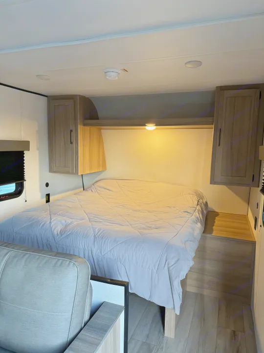 Inside an RV rental: a cozy bedroom with a large grey bed, overhead cabinets, and soft reading lights.. Colman 23B 2025