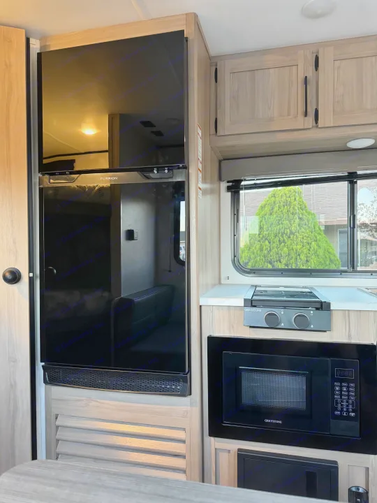 An RV rental interior showcasing a black Furrion fridge/TV, Greystone stovetop/microwave, and light wood cabinetry.. Colman 23B 2025