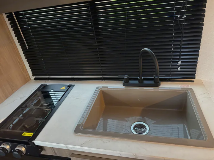 Compact RV rental kitchen with a two-burner stove, gray sink, black faucet, and horizontal window blinds.. Colman 23B 2025