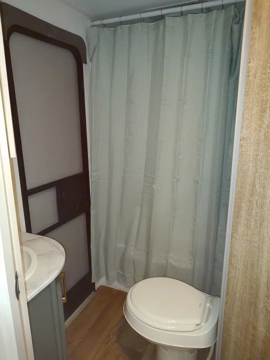 RV rental bathroom: compact toilet, light patterned shower curtain, sink vanity, and wood-look floor.. Heartland RVs Eddie Bauer 261BHC 2024