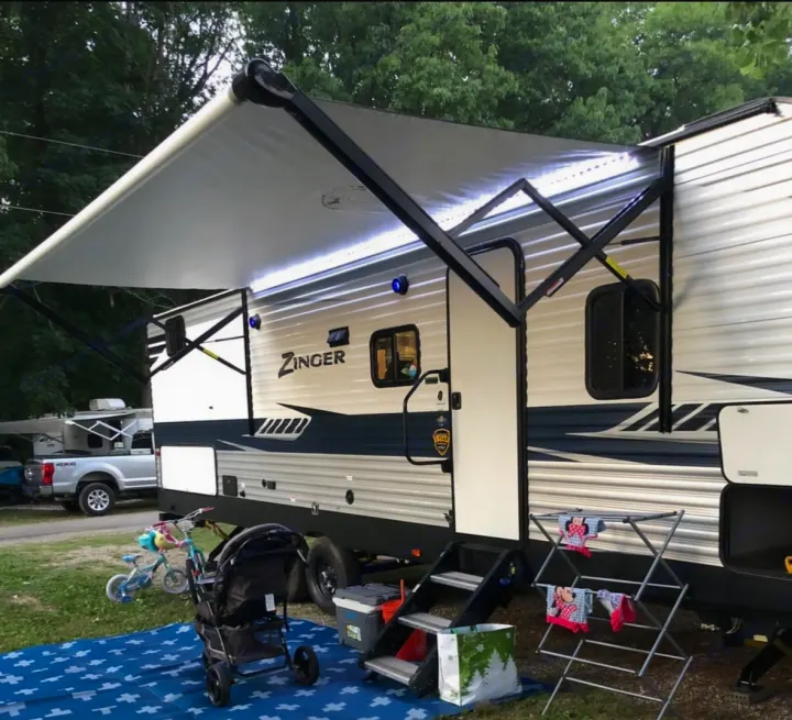 Spacious RV rental with illuminated awning set up at a campground. Features include outdoor mat, stroller, and clothesline.. Crossroads RV Zinger 2020