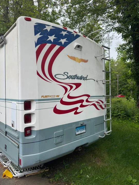 Rear of a Fleetwood Southwind RV, featuring a patriotic flag decal and ladder. This RV rental is parked in a green, wooded setting.. Fleetwood Southwind 36T 2000