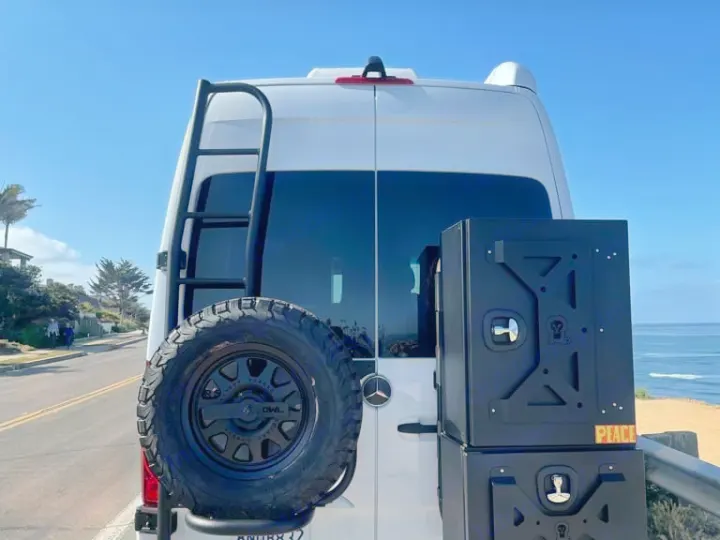 RV rental Sprinter van with rear ladder, spare tire, and storage boxes, parked by the ocean.. Airstream Interstate 2020