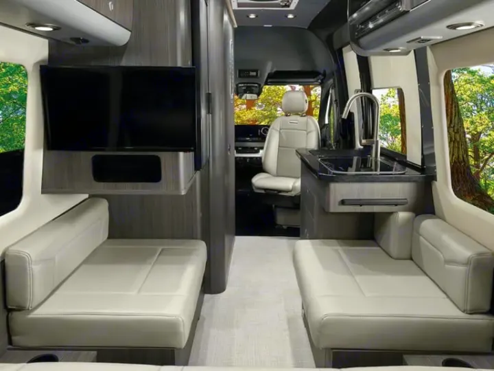 Spacious RV rental interior with side-facing benches, a wall-mounted TV, and compact kitchenette.. Airstream Interstate 2020