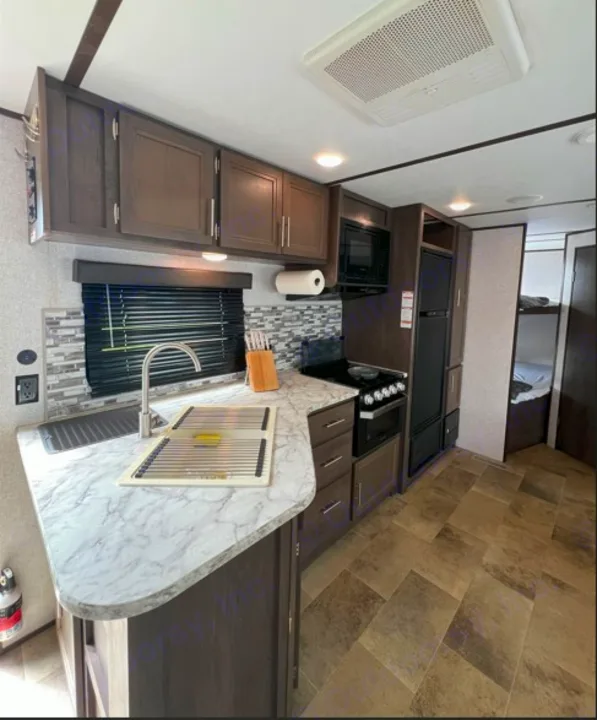 This RV rental kitchen features modern dark wood cabinets, marble-look counters, full appliances, and a sink. Bunks visible.. Dutchmen Coleman Lantern LT 2020