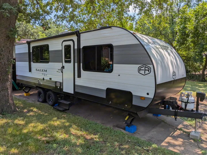 A white, grey, and black Forest River Salem FSX travel trailer parked on a driveway under green trees. An ideal RV rental.. Forest River Salem FSX 266BHLE 2025