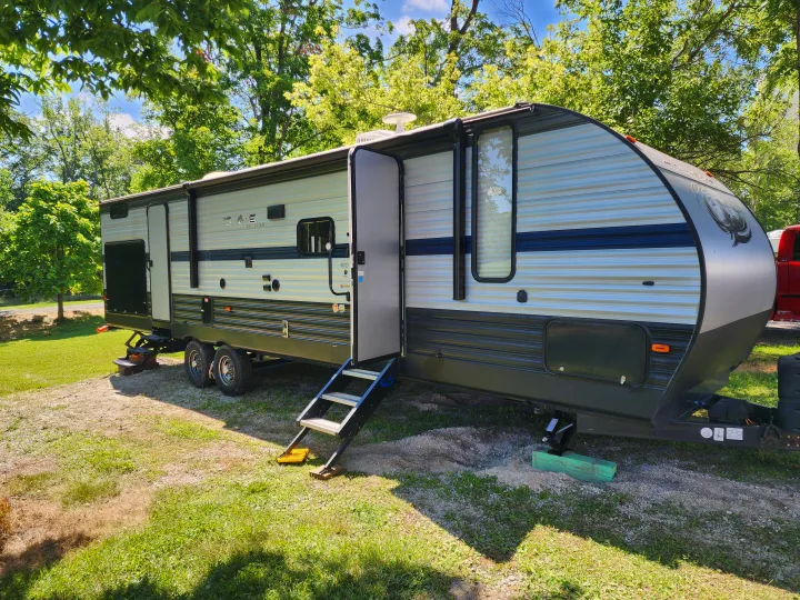 A modern grey and black travel trailer, an RV rental, parked on gravel and grass at a sunny campsite among trees.. Forest River Cherokee 2019