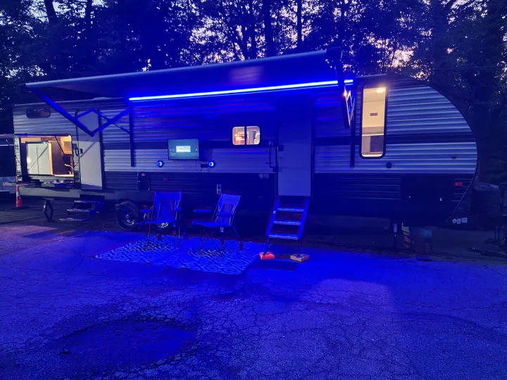 Evening view of a modern **rv rental** set up for outdoor living with bright blue awning lights, outdoor TV, and chairs.. Forest River Cherokee 2019