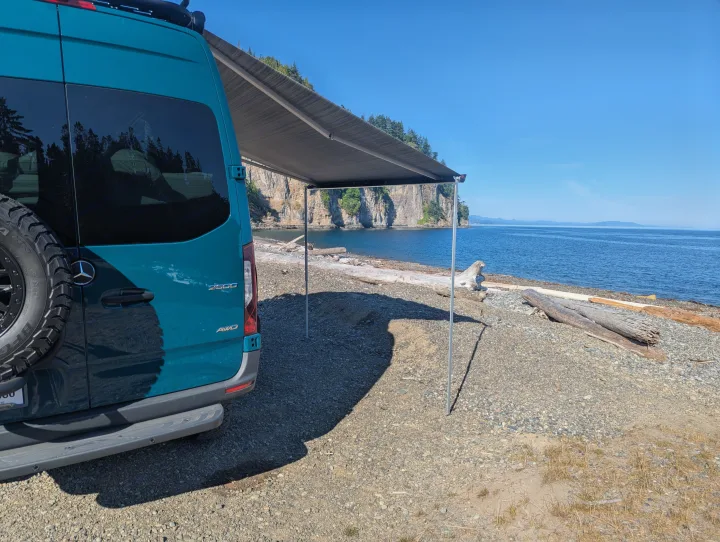 Teal RV rental with awning extended on a rocky beach by blue water & cliffs. Perfect for adventure!. Mercedes-Benz Sprinter 2025