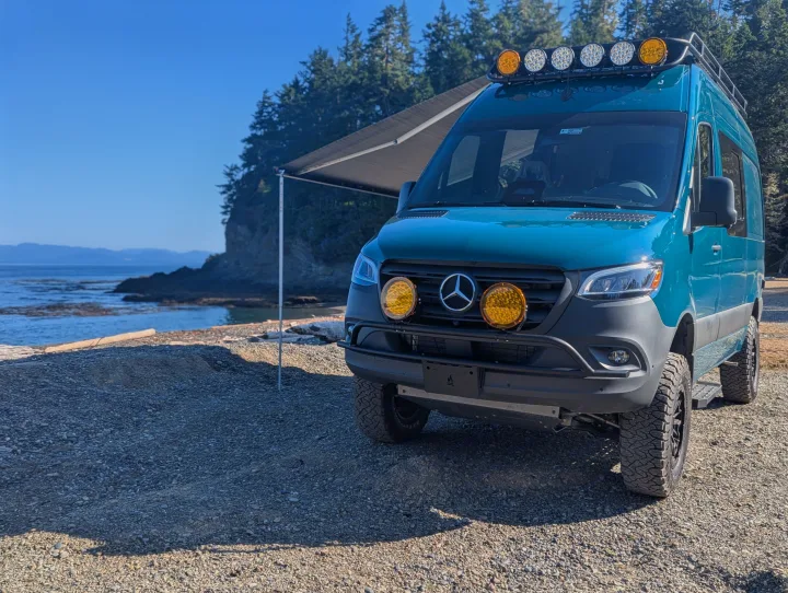 A teal Mercedes-Benz Sprinter camper van, an ideal rv rental, is parked on a rocky coast with an awning extended, facing the ocean.. Mercedes-Benz Sprinter 2025