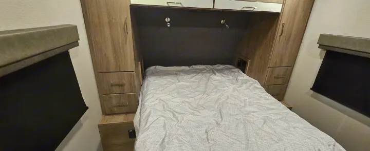 RV rental bedroom: a bed with white bedding, wooden side cabinets, overhead storage, and window blinds on either side.. Grand Design Imagine 2021