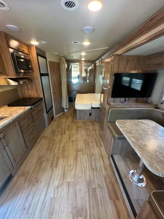 Spacious RV rental interior with kitchen, dining area, and bedroom featuring a bed and TV. Light wood-look flooring throughout.. Entegra Coach Odyssey 2019