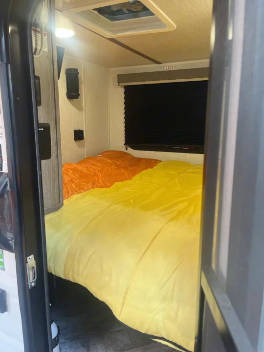 Cozy rv rental interior: a bed with vibrant orange and yellow bedding, and an EXIT sign above the window.. Sunset Park RV SunRay 2025