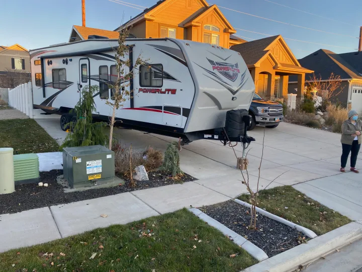 A large white travel trailer, possibly an RV rental, is parked in a residential driveway. A pickup truck and a woman are also visible.. Pacific Coachworks Powerlite 2017