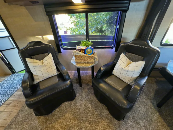 An RV rental's cozy seating area with two black chairs, a side table, s'mores supplies, and a window view.. Pacific Coachworks Powerlite 2017