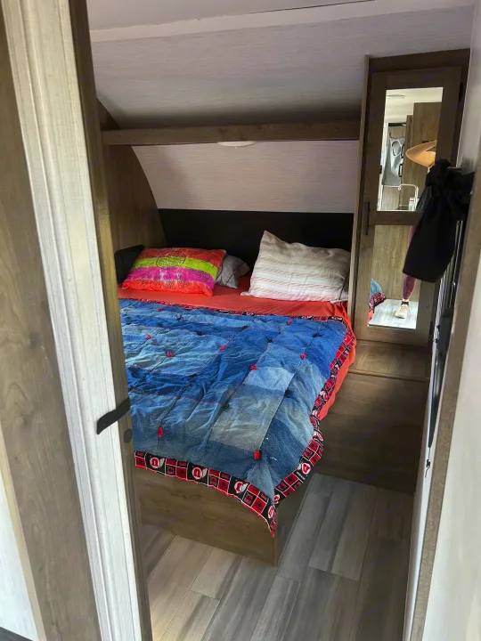 RV rental bedroom with a unique denim quilt, colorful pillows, and a mirror reflecting the adjacent kitchen.. Heartland RVs Pioneer 2022