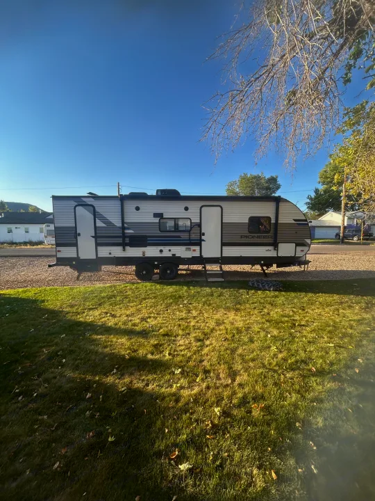 A white and gray Pioneer travel trailer RV rental is parked on a gravel lot with green grass in the foreground under a clear blue sky.. Heartland RVs Pioneer 2022