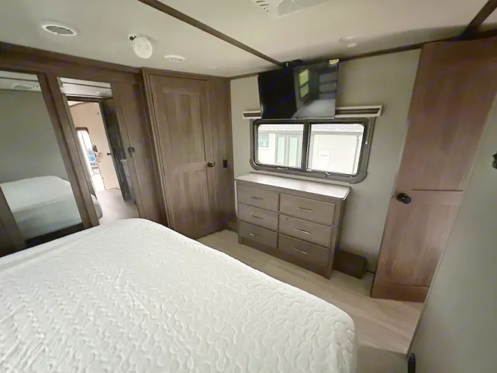 Inside an RV rental: a compact bedroom featuring a white bed, dresser, TV, and reflective closet door.. Grand Design Solitude 2022
