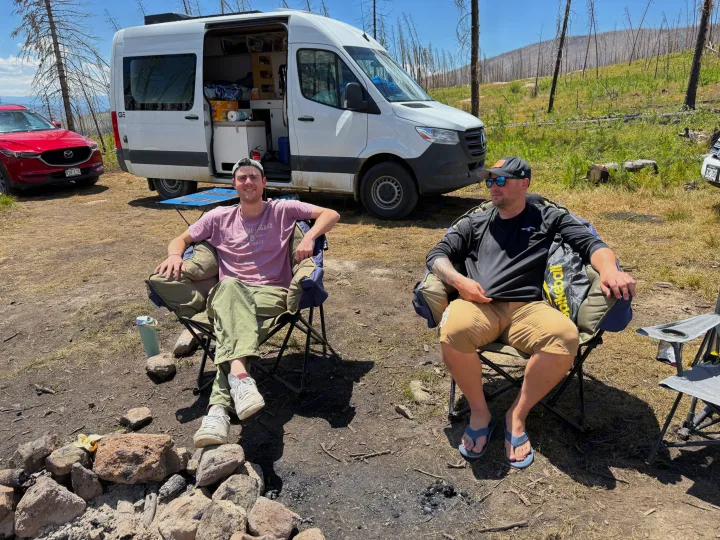 Two men relaxing at a campsite with an RV rental and a red SUV, near a fire pit in a scenic, treed area.. Mercedes-Benz Sprinter 2024