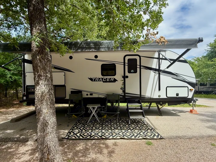 A Primetime Tracer RV rental set up at a spacious campsite with an extended awning, outdoor rug, camping chairs, and table for comfort.. Forest River Tracer 2022