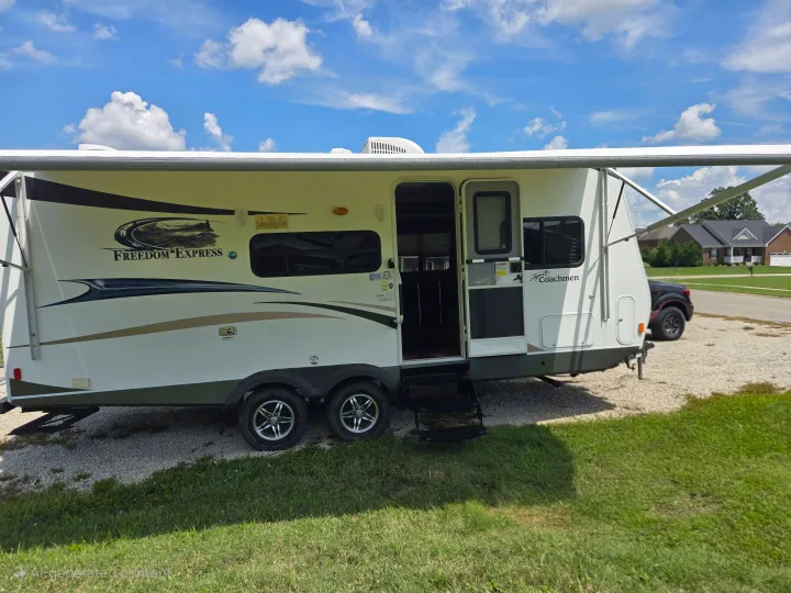View of a white "Freedom Express" travel trailer RV rental, with extended awning & open door, parked on gravel under a sunny sky.. Coachmen Freedom express ltz 2014
