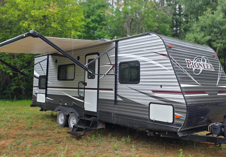 This grey Pioneer RV rental travel trailer with an extended awning is parked in a grassy area amidst trees.. Heartland RVs Pioneer 2017
