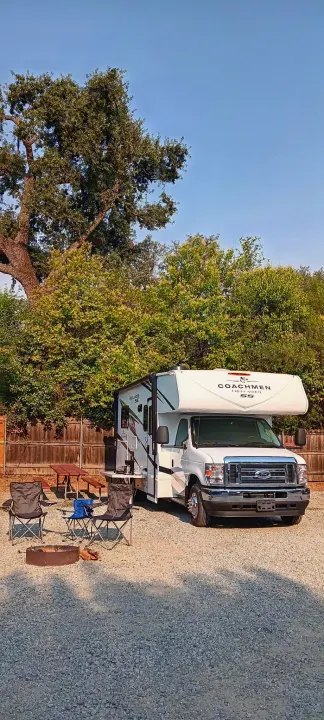 White Coachmen RV rental parked at a gravel campsite. Features a fire pit, folding chairs, & picnic table for outdoor fun.. Freelander 21QSS 2025