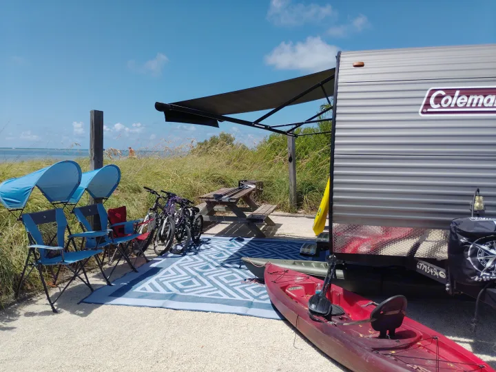 Coleman RV rental parked at a sunny beach campsite, complete with awning, kayaks, bikes, chairs, and outdoor rug.. Dutchmen 17B 2024