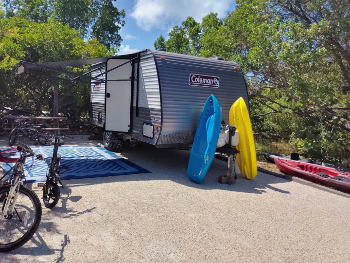 RV rental campsite setup: A grey Coleman travel trailer with awning, bikes, and colorful kayaks on a sunny gravel lot.. Dutchmen 17B 2024