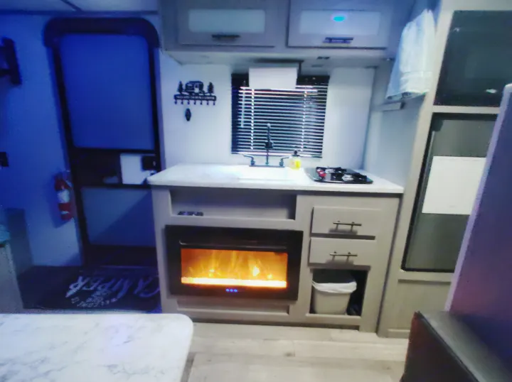 Inside an RV rental: a cozy kitchen with sink, stove, and electric fireplace. Perfect compact living for road trips.. Dutchmen 17B 2024