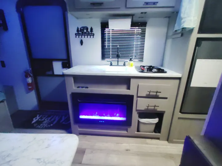 Modern RV rental interior featuring a compact kitchen with sink, stove, and a vibrant purple electric fireplace.. Dutchmen 17B 2024
