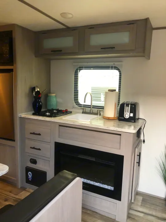 Modern RV rental kitchen featuring a compact sink, stovetop, refrigerator, and electric fireplace. Clean, light-toned interior.. Coleman Lantern 17R 2024