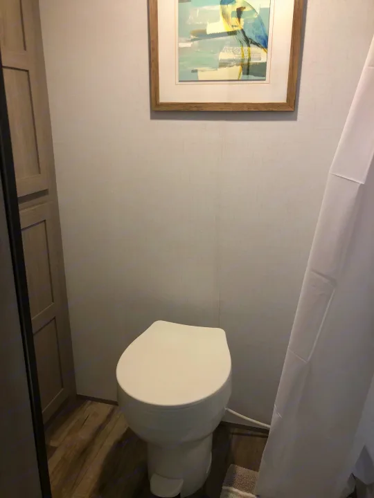 Compact RV rental bathroom showcasing a toilet, wood floor, white shower curtain, and wall art.. Coleman Lantern 17R 2024