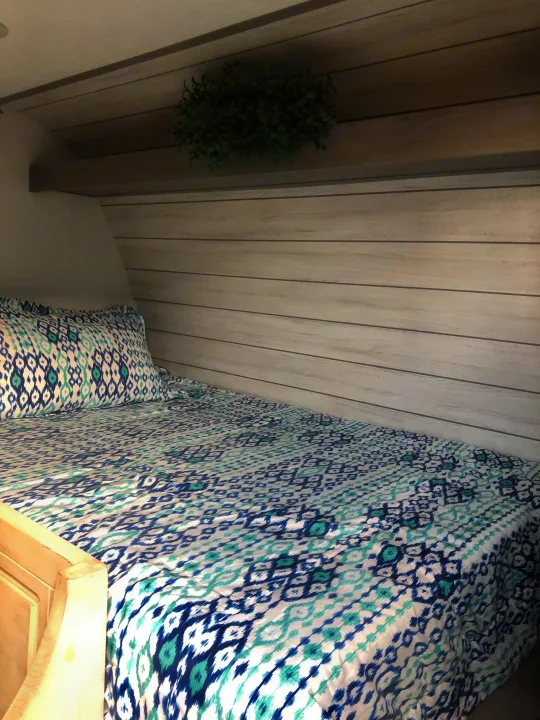 Cozy RV rental interior featuring a built-in bed with blue/teal patterned bedding, wood paneling, and a shelf with a plant.. Coleman Lantern 17R 2024