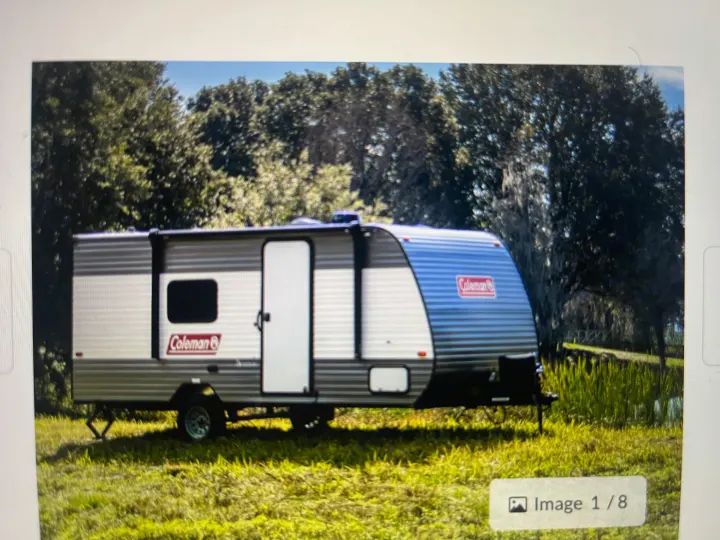 A silver and blue Coleman travel trailer RV rental is parked on green grass amidst trees, ready for adventure.. Coleman Lantern 17R 2024