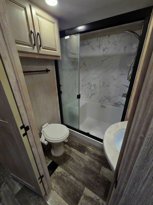 Clean RV rental bathroom with a compact design, featuring a toilet, walk-in glass shower, sink, and light wood textures.. Forest River Sandpiper 2023