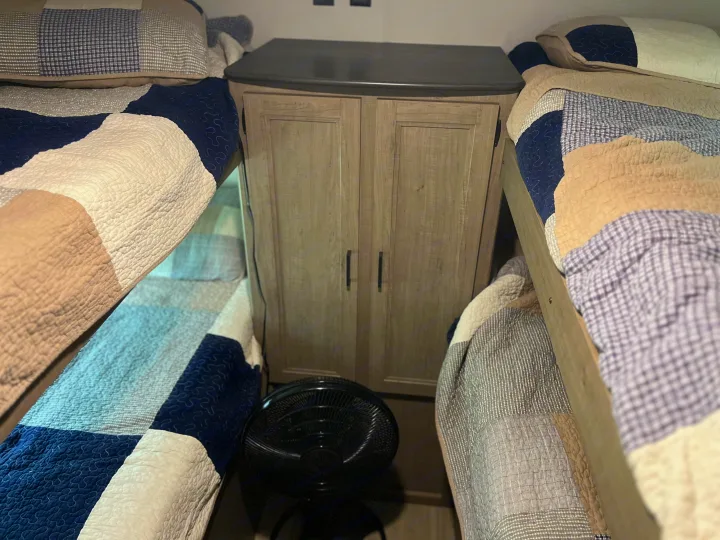 An RV rental interior shows two cozy bunk beds with patchwork quilts, a central storage cabinet, and a floor fan.. Keystone RV Hideout LHS 2020