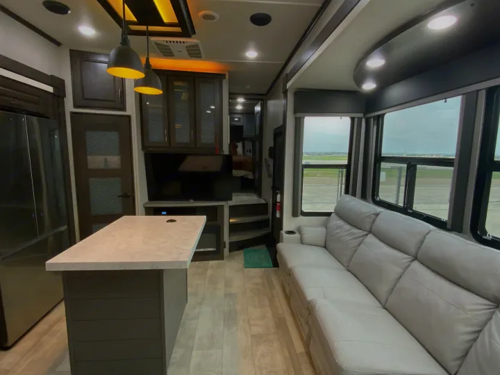 Spacious RV rental living area with a modern kitchen island, gray sofa, and bright windows showcasing the view.. Grand Design Momentum 2024