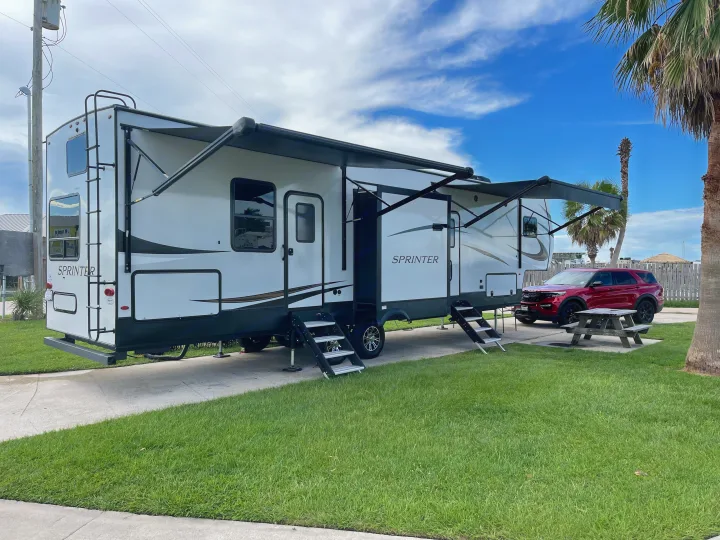Large white RV rental with extended awnings and steps parked at a sunny campsite with a red SUV, picnic table, and palm trees.. Keystone RV Sprinter 2022