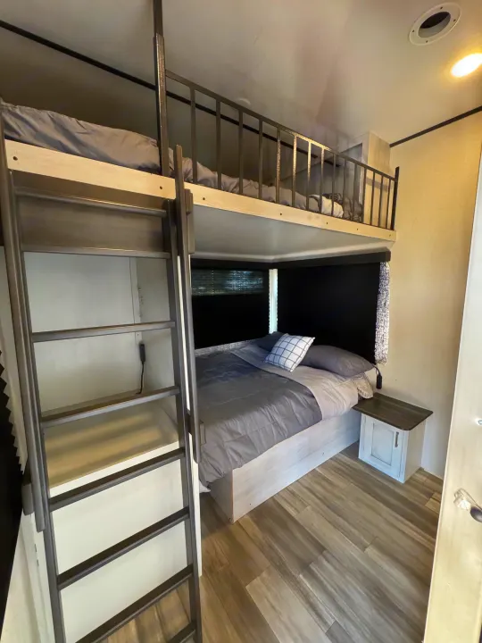 This RV rental bedroom features a lower bed with grey bedding, an upper bunk with a safety railing, and an access ladder. Black blinds & a nightstand are visible.. Keystone RV Sprinter 2022