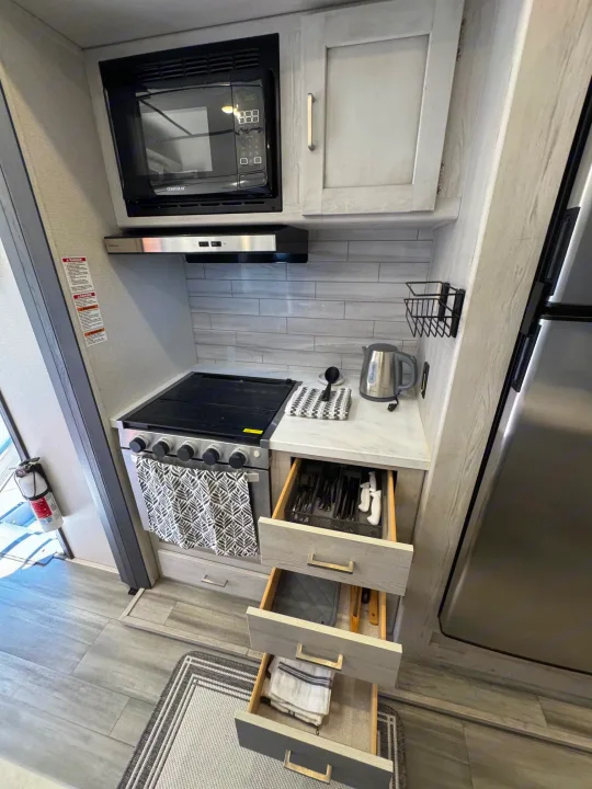 Clean, organized RV rental kitchen area: microwave, stove, open drawers of cutlery, kettle, and a visible fire extinguisher.. Keystone RV Sprinter 2022