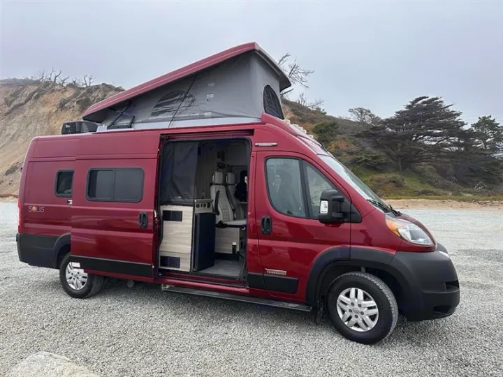 Red pop-top RV rental camper van with open side door revealing its interior, parked on gravel by a coastal hill.. Winnebago Winnebago 2023