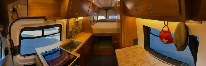 Modern camper van RV rental interior featuring a kitchen with sink and stove, plus a cozy bed area and wood cabinets.. Ford Transit 2018